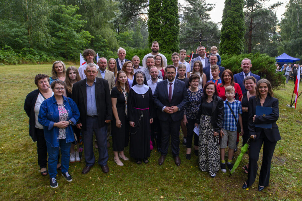 Grupa ludzi pozuje do zdjęcia na tle zielonego lasu. Mają różnorodne stroje, w tym eleganckie garnitury i sukienki. W oddali widać niebieski namiot oraz flagi.
