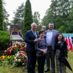 Grupa ludzi przed grobem z kwiatami czerwono-białymi. Tło to zielony park z drzewami. Po prawej flagi Polski.