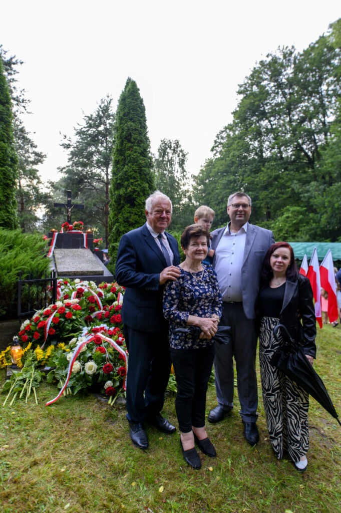 Grupa ludzi przed grobem z kwiatami czerwono-białymi. Tło to zielony park z drzewami. Po prawej flagi Polski.