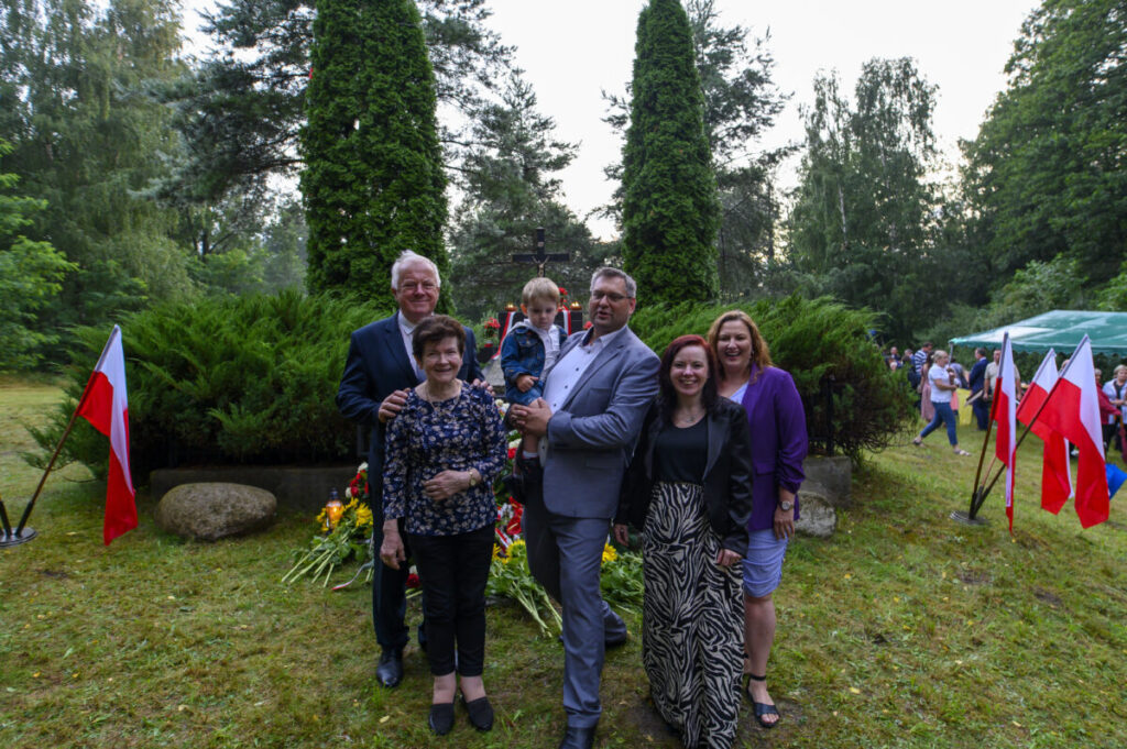 Grupa sześciu osób stoi przed zielenią i wysokimi krzewami. W tle widać gęsty las i tłum ludzi. Wokół powiewają polskie flagi.