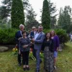 Grupa sześciu osób stoi przed zielenią i wysokimi krzewami. W tle widać gęsty las i tłum ludzi. Wokół powiewają polskie flagi.