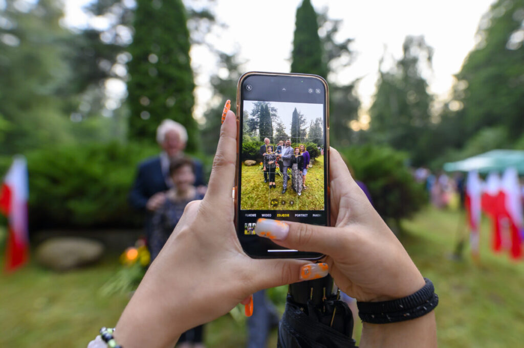 Telefon w rękach fotografuje grupę osób na tle zieleni. W tle widać drzewa i flagi. Kolory są jasne, dominują zieleń i biel.