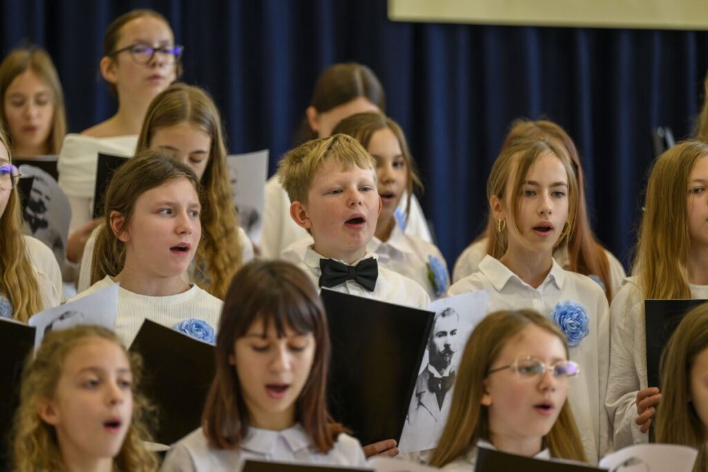 Grupa dzieci ubranych na biało śpiewa. Mają czarne teczki z nutami i niebieskie kwiaty. Tło stanowi ciemna zasłona.