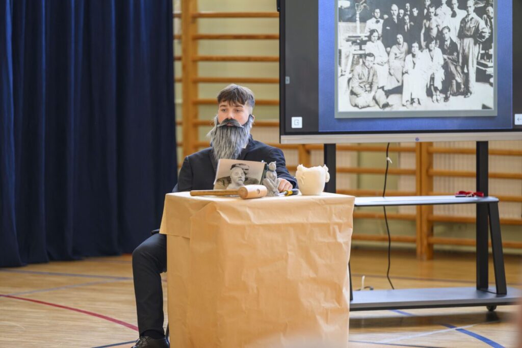 Mężczyzna w przebraniu siedzi przy stole pokrytym papierem. Na ekranie za nim widać czarno-białą fotografię grupową. Tło jest granatowe z drewnianymi elementami.