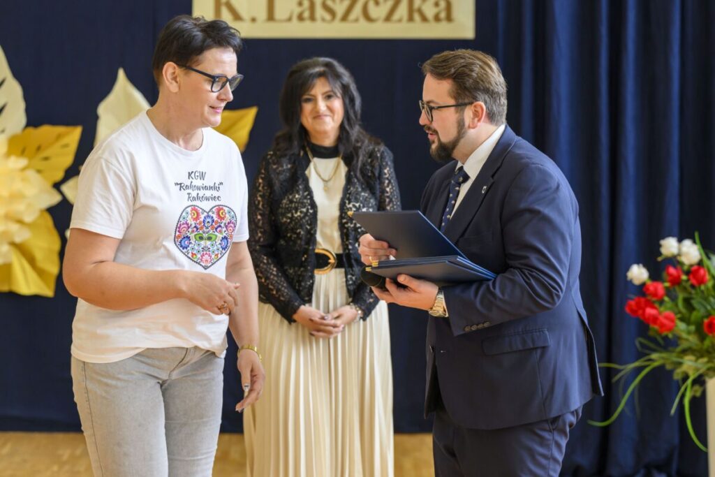 Trzy osoby stoją w jasnym pokoju; kobieta w białej koszulce, mężczyzna w garniturze wręcza jej dokument. Tło ciemnoniebieskie z kwiatami.