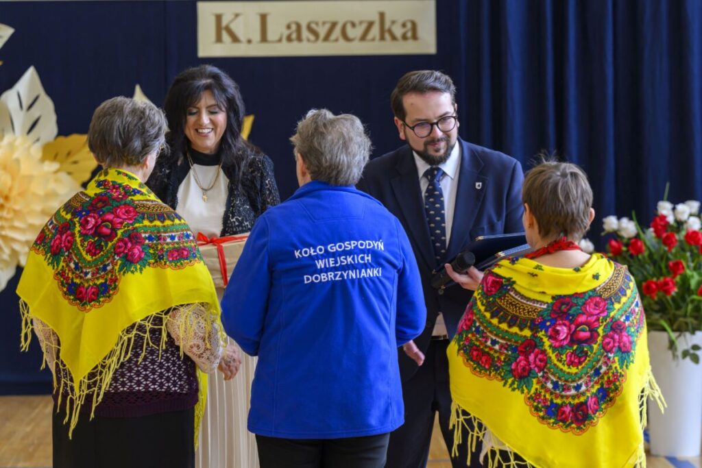 Na zdjęciu dwie kobiety w żółtych chustach i kobieta oraz mężczyzna w eleganckich strojach. Tło jest ciemnoniebieskie z napisem. Widać czerwone kwiaty w wazonach.