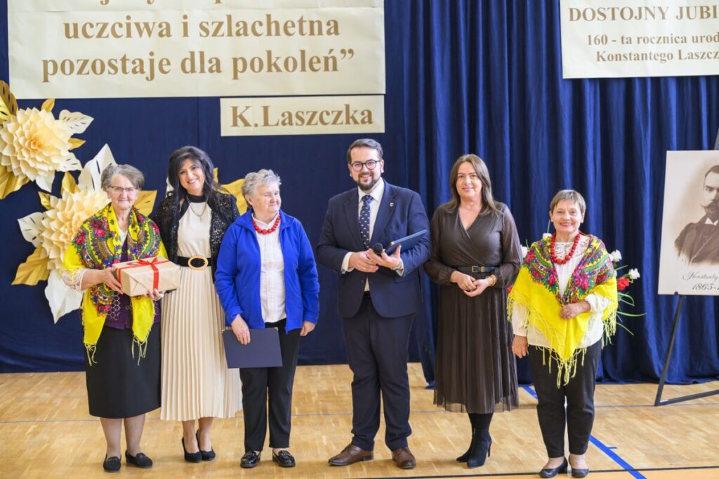 Grupa sześciu osób stoi przed niebieską kurtyną. Dwie kobiety mają kolorowe szale. Tło zdobią papierowe kwiaty i plakaty.