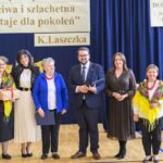 Grupa sześciu osób stoi przed niebieską kurtyną. Dwie kobiety mają kolorowe szale. Tło zdobią papierowe kwiaty i plakaty.