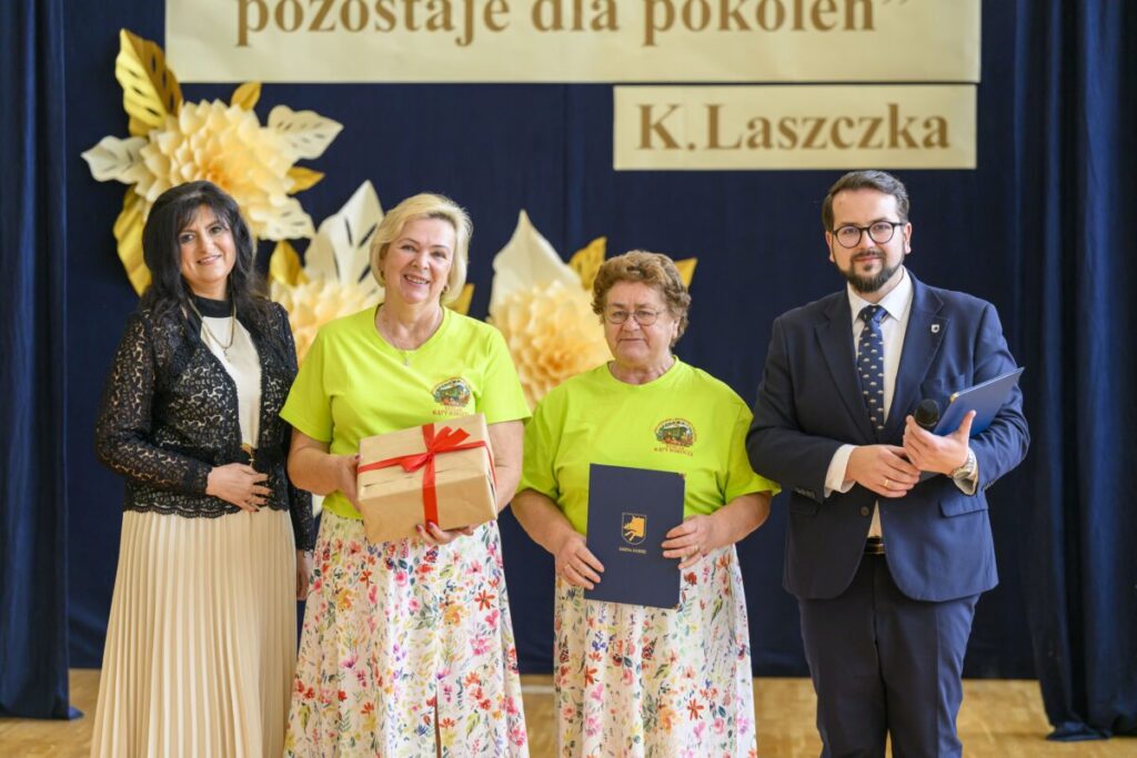 Na zdjęciu cztery osoby na tle dekoracji z żółtych i białych kwiatów. Dwie kobiety w zielonych koszulkach trzymają prezenty. Mężczyzna i kobieta są elegancko ubrani.