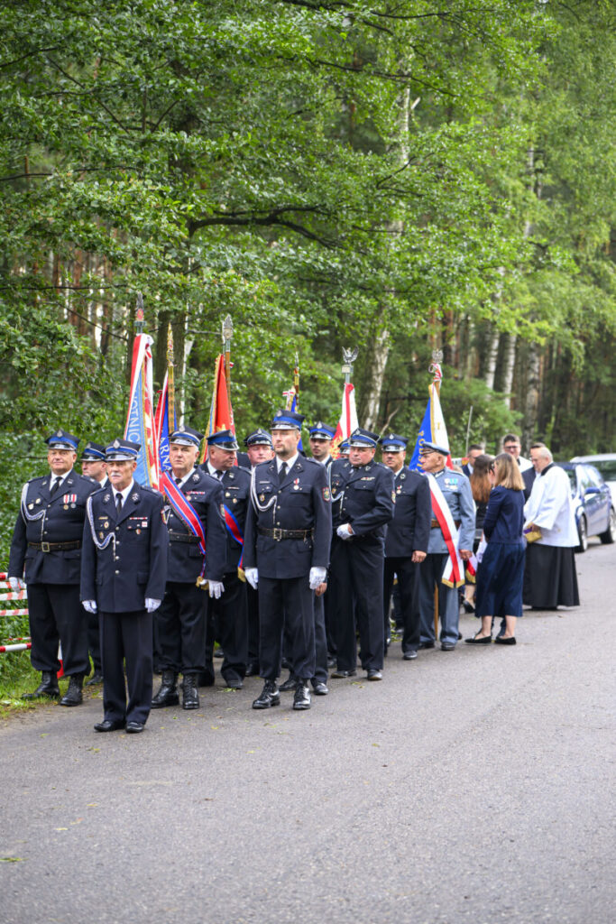 Grupa strażaków w mundurach stoi w szeregu z flagami. W tle zielone drzewa i zaparkowany samochód. Uroczysta atmosfera.