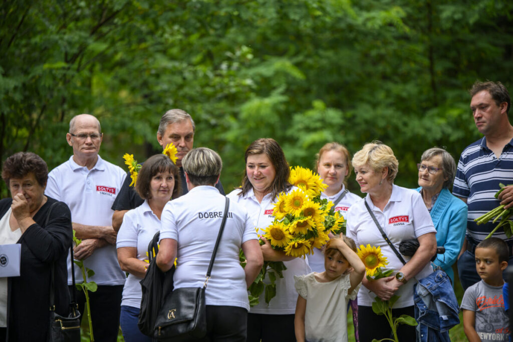 Grupa osób w białych koszulkach z napisami stoi wśród drzew. Osoby trzymają bukiety słoneczników. Tło jest zielone, leśne.