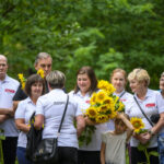 Grupa osób w białych koszulkach z napisami stoi wśród drzew. Osoby trzymają bukiety słoneczników. Tło jest zielone, leśne.