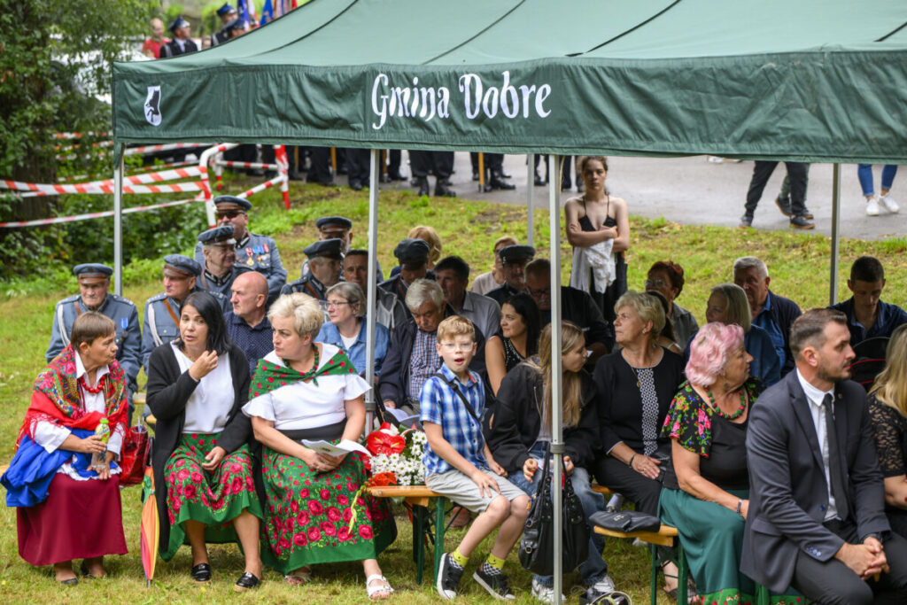 Grupa ludzi siedzi pod zielonym namiotem w parku. Niektórzy noszą kolorowe regionalne stroje, a w tle widać trawę. Napis na namiocie: Gmina Dobre.