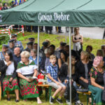 Grupa ludzi siedzi pod zielonym namiotem w parku. Niektórzy noszą kolorowe regionalne stroje, a w tle widać trawę. Napis na namiocie: Gmina Dobre.