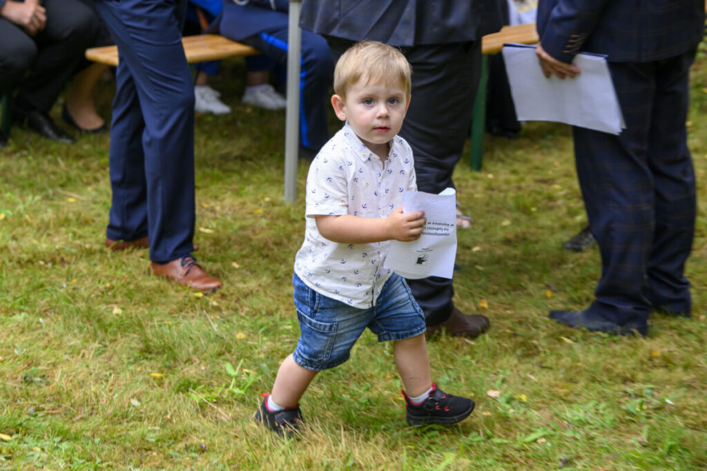 Na zdjęciu jest mały chłopiec w białej koszuli i jeansowych spodenkach, trzyma kartkę. Jest na trawie, otoczony przez osoby w ciemnych garniturach. Tło jest rozmyte, sugerujące plenerowe wydarzenie.
