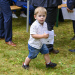 Na zdjęciu jest mały chłopiec w białej koszuli i jeansowych spodenkach, trzyma kartkę. Jest na trawie, otoczony przez osoby w ciemnych garniturach. Tło jest rozmyte, sugerujące plenerowe wydarzenie.