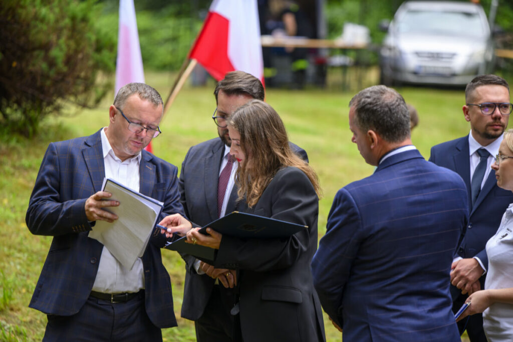 Grupa osób w eleganckiej odzieży stoi na zielonej trawie. Obok nich widać flagi Polski. Tło to samochód i drzewa.