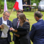 Grupa osób w eleganckiej odzieży stoi na zielonej trawie. Obok nich widać flagi Polski. Tło to samochód i drzewa.