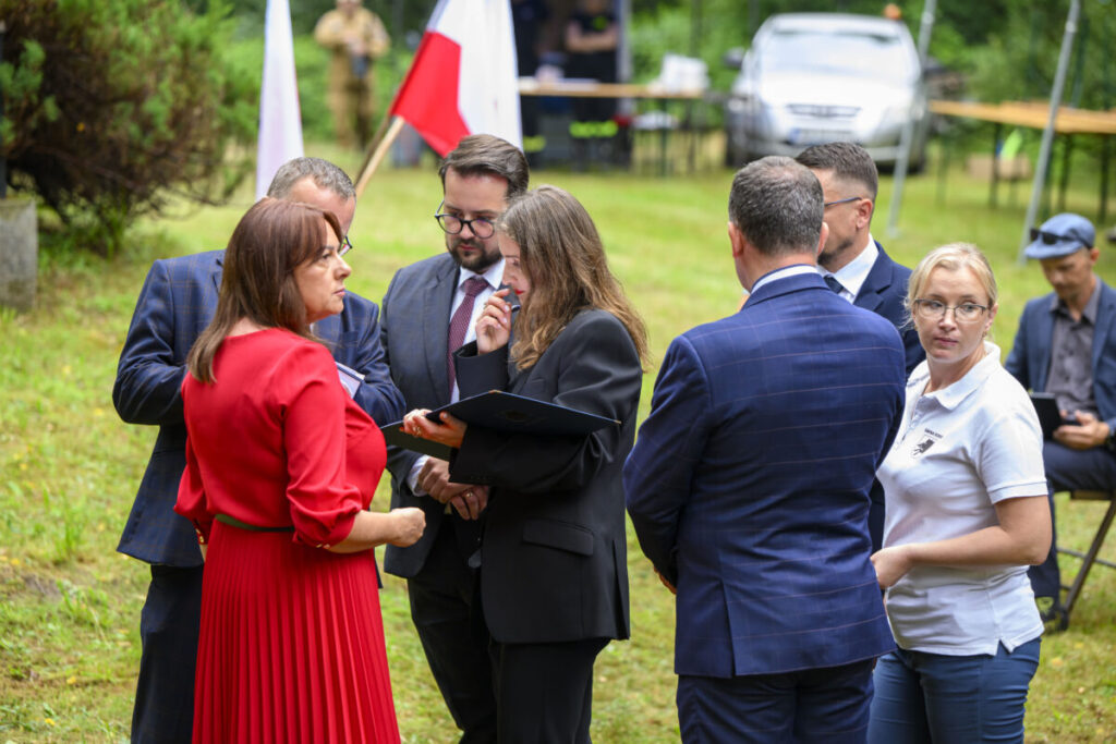 Grupa osób w eleganckich ubraniach rozmawia na zewnątrz. Kobieta w czerwieni, obok biało-czerwone flagi. W tle trawiasty teren i samochód.