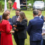 Grupa osób w eleganckich ubraniach rozmawia na zewnątrz. Kobieta w czerwieni, obok biało-czerwone flagi. W tle trawiasty teren i samochód.