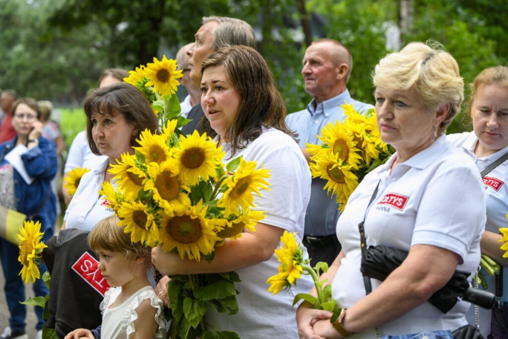 Grupa ludzi w białych koszulkach trzyma bukiety słoneczników. W tle zielone liście drzew. Jasne, letnie kolory dominują.
