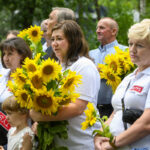 Grupa ludzi w białych koszulkach trzyma bukiety słoneczników. W tle zielone liście drzew. Jasne, letnie kolory dominują.