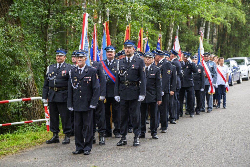 Grupa strażaków w mundurach i czapkach stoi na drodze, w tle zieleń lasu. Noszą medale i trzymają różnobarwne flagi. Tło jest zielone i spokojne.