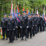 Grupa strażaków w mundurach i czapkach stoi na drodze, w tle zieleń lasu. Noszą medale i trzymają różnobarwne flagi. Tło jest zielone i spokojne.