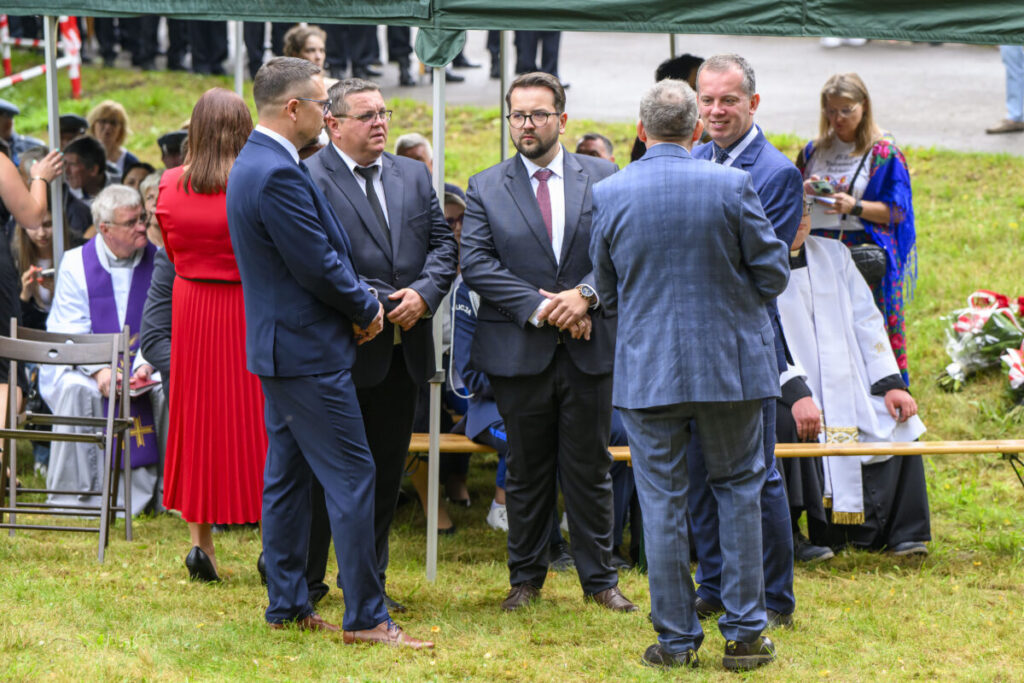 Na zdjęciu kilku mężczyzn w garniturach rozmawia na zielonej trawie. W tle namiot, osoby siedzące, w tym duchowny w białej albie. Kobieta w czerwonej sukni stoi, obok kobieta z telefonem w tle.