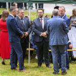 Na zdjęciu kilku mężczyzn w garniturach rozmawia na zielonej trawie. W tle namiot, osoby siedzące, w tym duchowny w białej albie. Kobieta w czerwonej sukni stoi, obok kobieta z telefonem w tle.