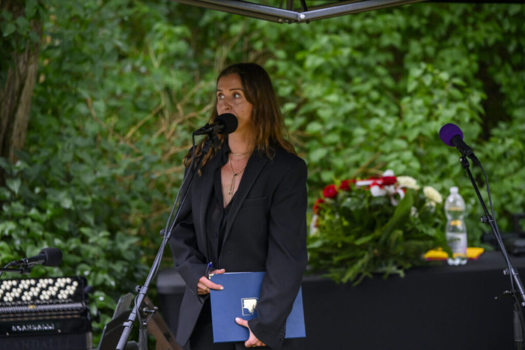 Osoba w czarnym garniturze przemawia do mikrofonu. Zielone tło z krzewami. Na stole obok leży bukiet kwiatów.