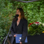 Osoba w czarnym garniturze przemawia do mikrofonu. Zielone tło z krzewami. Na stole obok leży bukiet kwiatów.