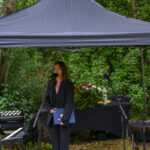 Kobieta w czerni przemawia pod czarnym namiotem w ogrodzie. W tle zielone liście i stół z kwiatami. Obok keyboard.