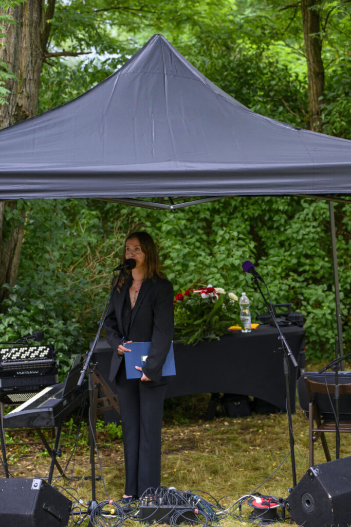 Kobieta w czerni przemawia pod czarnym namiotem w ogrodzie. W tle zielone liście i stół z kwiatami. Obok keyboard.