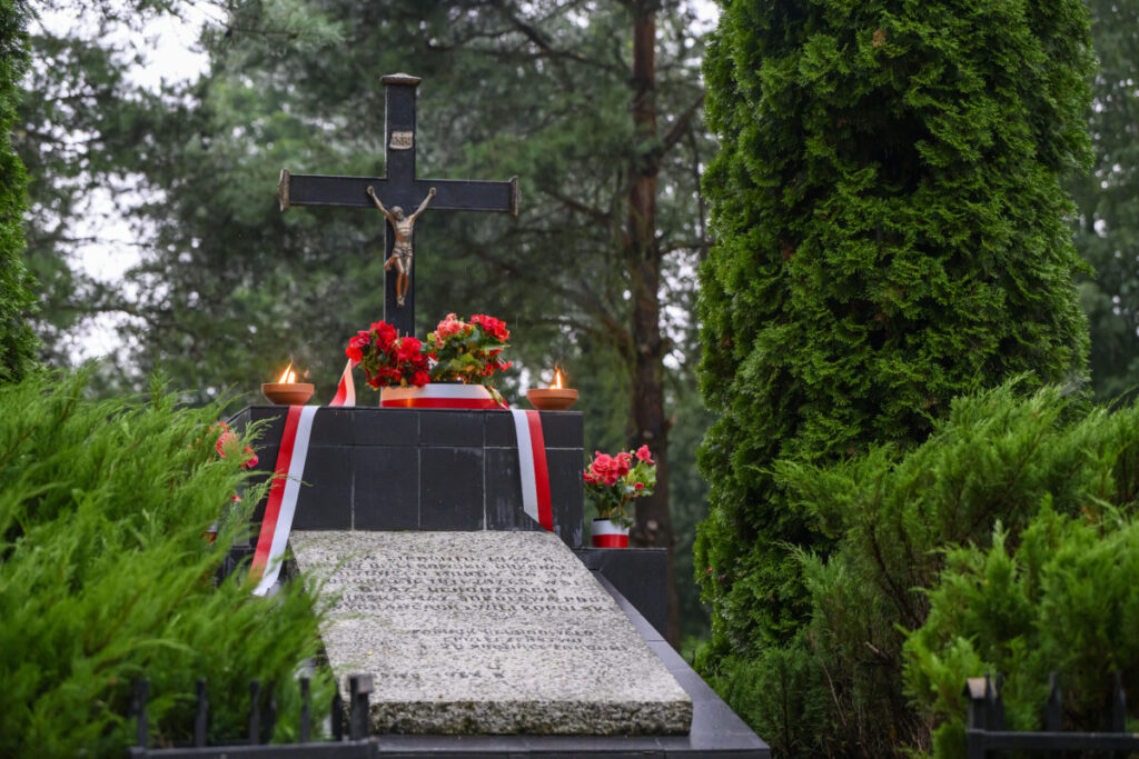 Grób z krzyżem i Chrystusem pośród zielonych drzew. Czerwono-białe kwiaty i wstęga dekorują miejsce. Pochmurne niebo w tle.