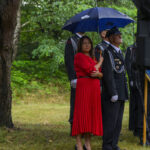 Na zdjęciu kobieta w czerwonej sukni pod niebieskim parasolem z mężczyzną w mundurze. Tło to zielony las. Atmosfera jest deszczowa.