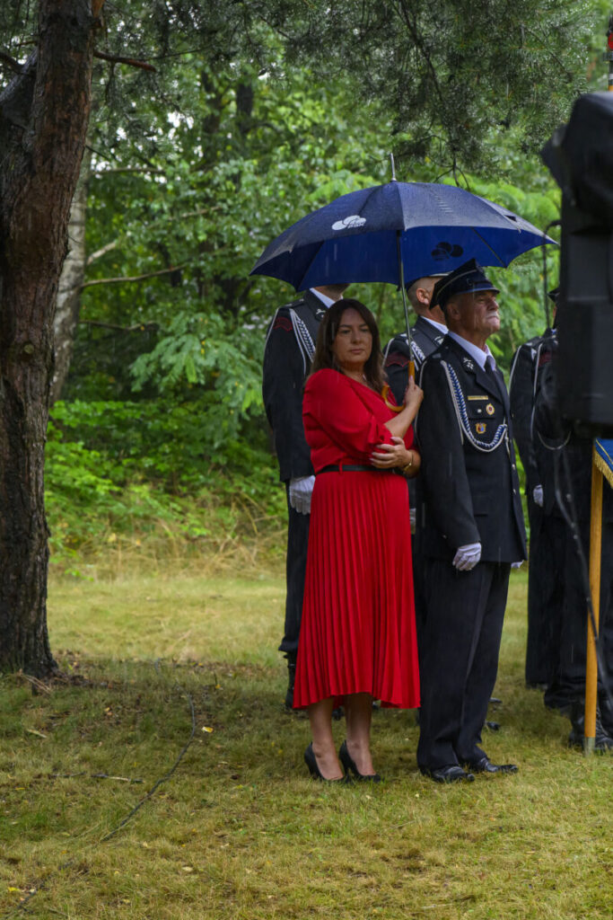 Na zdjęciu kobieta w czerwonej sukni pod niebieskim parasolem z mężczyzną w mundurze. Tło to zielony las. Atmosfera jest deszczowa.
