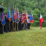 Uroczystość z udziałem osób w mundurach trzymających kolorowe sztandary na tle zieleni. Widoczne są też biało-czerwone flagi. Tło stanowią drzewa i trawa.