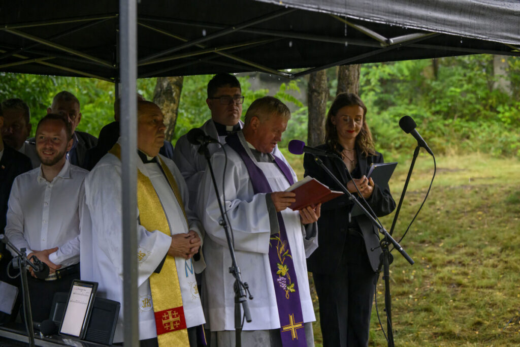 Grupa ludzi pod czarnym namiotem; duchowni w białych szatach, mikrofony z przodu. W tle zielona roślinność.