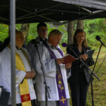 Grupa ludzi pod czarnym namiotem; duchowni w białych szatach, mikrofony z przodu. W tle zielona roślinność.
