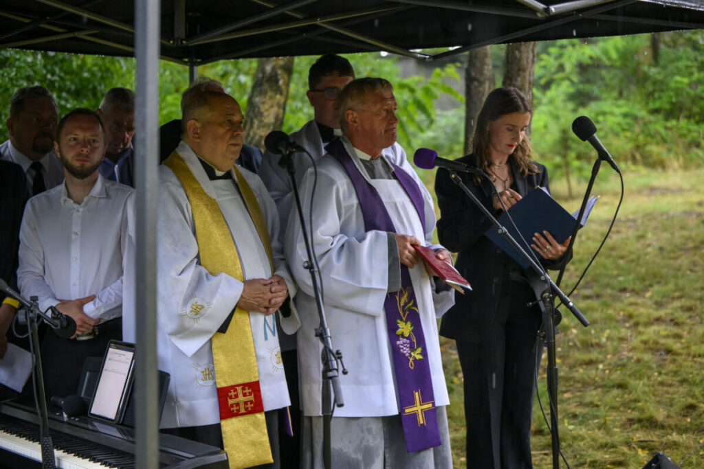 Na zdjęciu kilku księży w białych i ozdobnych szatach, obok kobieta z książką. Tło zielone, z drzewami. Mają mikrofony pod namiotem.