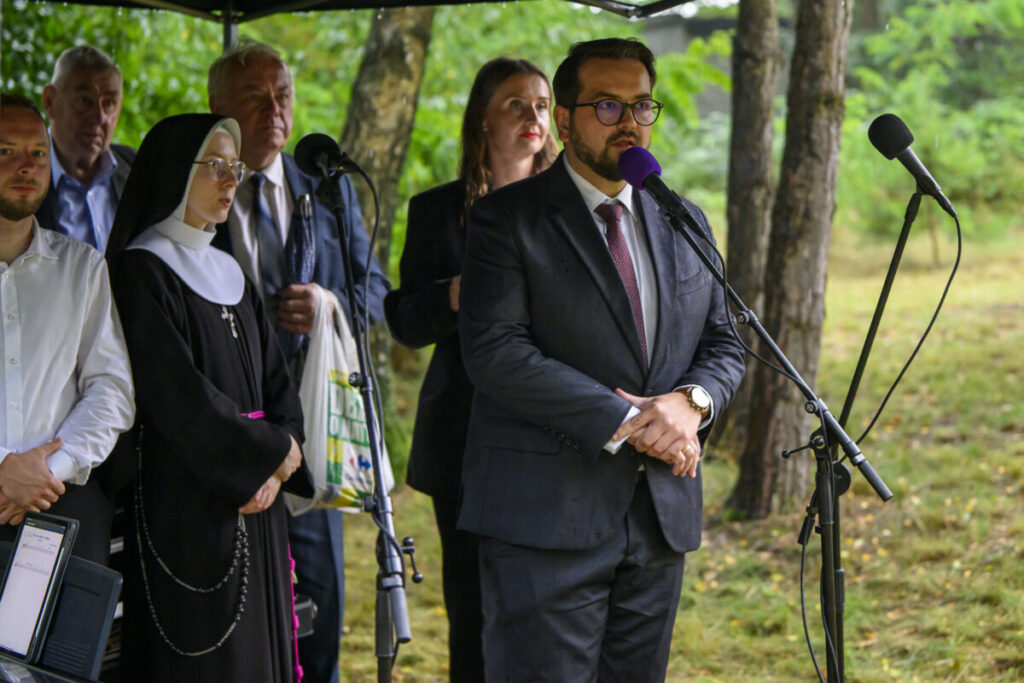 Grupa osób stoi przy mikrofonach, w tym zakonnica i mężczyzna w garniturze. Tło to zielone drzewa. Scena plenerowa.