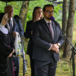 Grupa osób stoi przy mikrofonach, w tym zakonnica i mężczyzna w garniturze. Tło to zielone drzewa. Scena plenerowa.