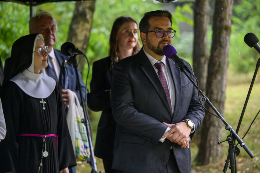 Zdjęcie przedstawia mężczyznę w garniturze przemawiającego do mikrofonu. Obok stoi zakonnica, w tle drzewa. Kolory dominujące to zieleń i szarość.