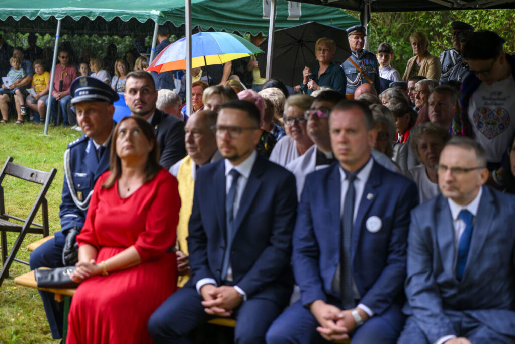 Grupa ludzi siedzi pod zielonym namiotem; na pierwszym planie osoby elegancko ubrane, w tle widoczne kolorowe parasole.