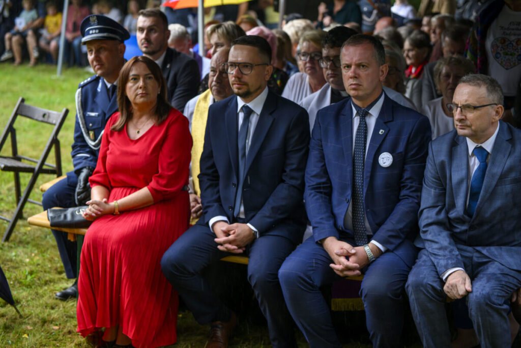 Na zdjęciu widoczni są ludzie siedzący na zewnątrz w eleganckim stroju, zielona trawa dookoła. Mężczyzna w mundurze, kobieta w czerwonej sukience. Grupa osób siedzi na ławce, tło rozmyte.