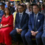 Na zdjęciu widoczni są ludzie siedzący na zewnątrz w eleganckim stroju, zielona trawa dookoła. Mężczyzna w mundurze, kobieta w czerwonej sukience. Grupa osób siedzi na ławce, tło rozmyte.