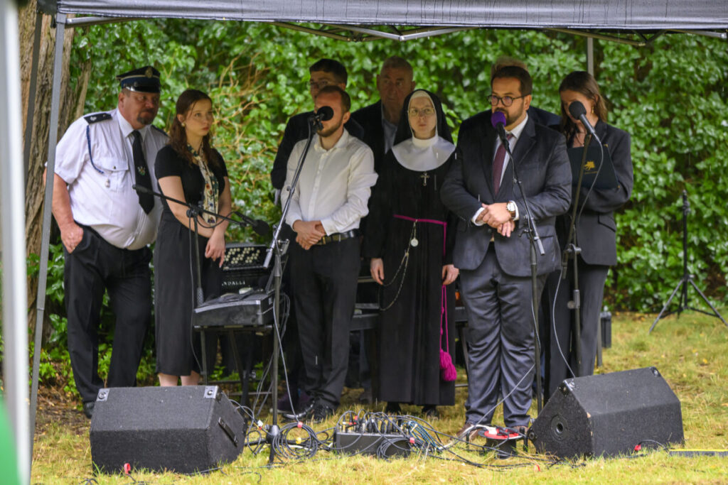 Grupa osób stoi pod namiotem na tle zieleni, przemawiają do mikrofonów. Są ubrani w formalne stroje. Na trawie leżą kable i sprzęt nagłaśniający.