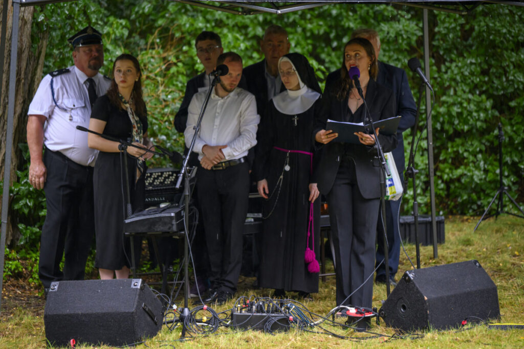 Grupa osób, w tym zakonnica, stoi na scenie pod zadaszeniem. Mikrofony i sprzęt audio na trawie. Zielone tło z drzewami.
