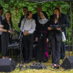 Grupa osób, w tym zakonnica, stoi na scenie pod zadaszeniem. Mikrofony i sprzęt audio na trawie. Zielone tło z drzewami.